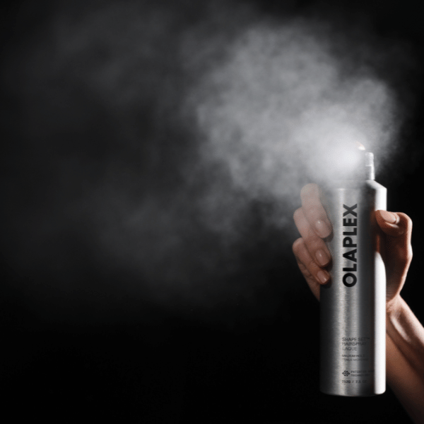Hand sprueht OLAPLEX Shape Set Hairspray mit sichtbar feinem Spruehnebel vor dunklem Hintergrund