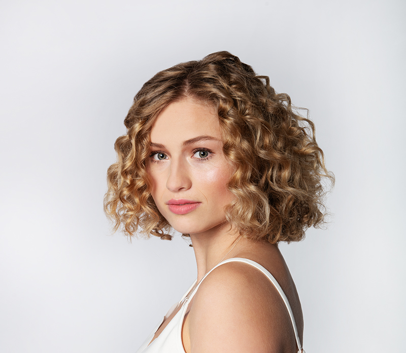 frau-mit-blonden-naturlocken-und-schulterlangem-haar-blickt-in-die-kamera–natuerlicher-beauty-look-mit-heller-haut-und-zarten-make-up