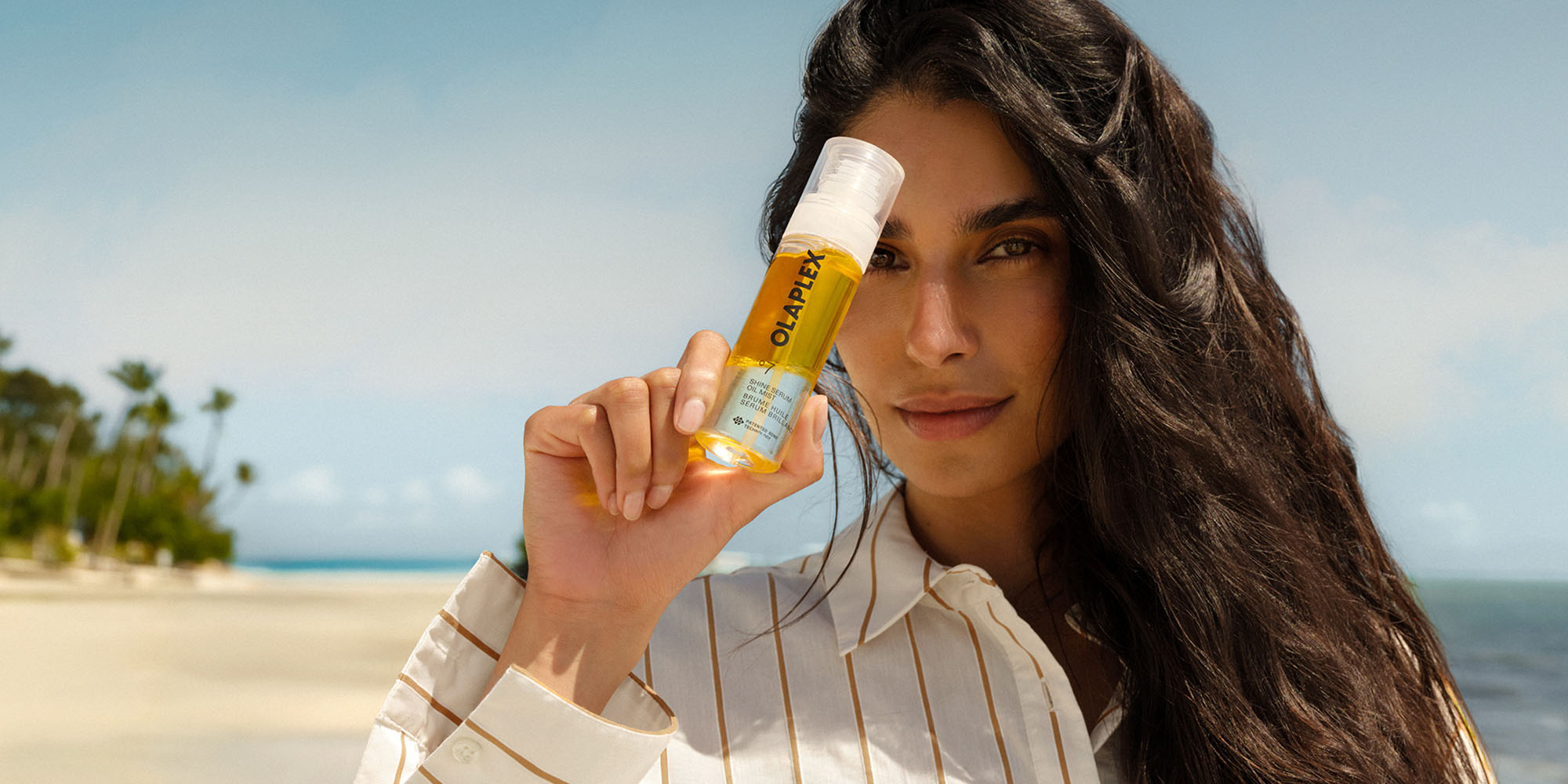 Frau mit langem dunklem Haar haelt eine Flasche OLAPLEX No. 7 Shine Serum neben ihr Gesicht am Strand