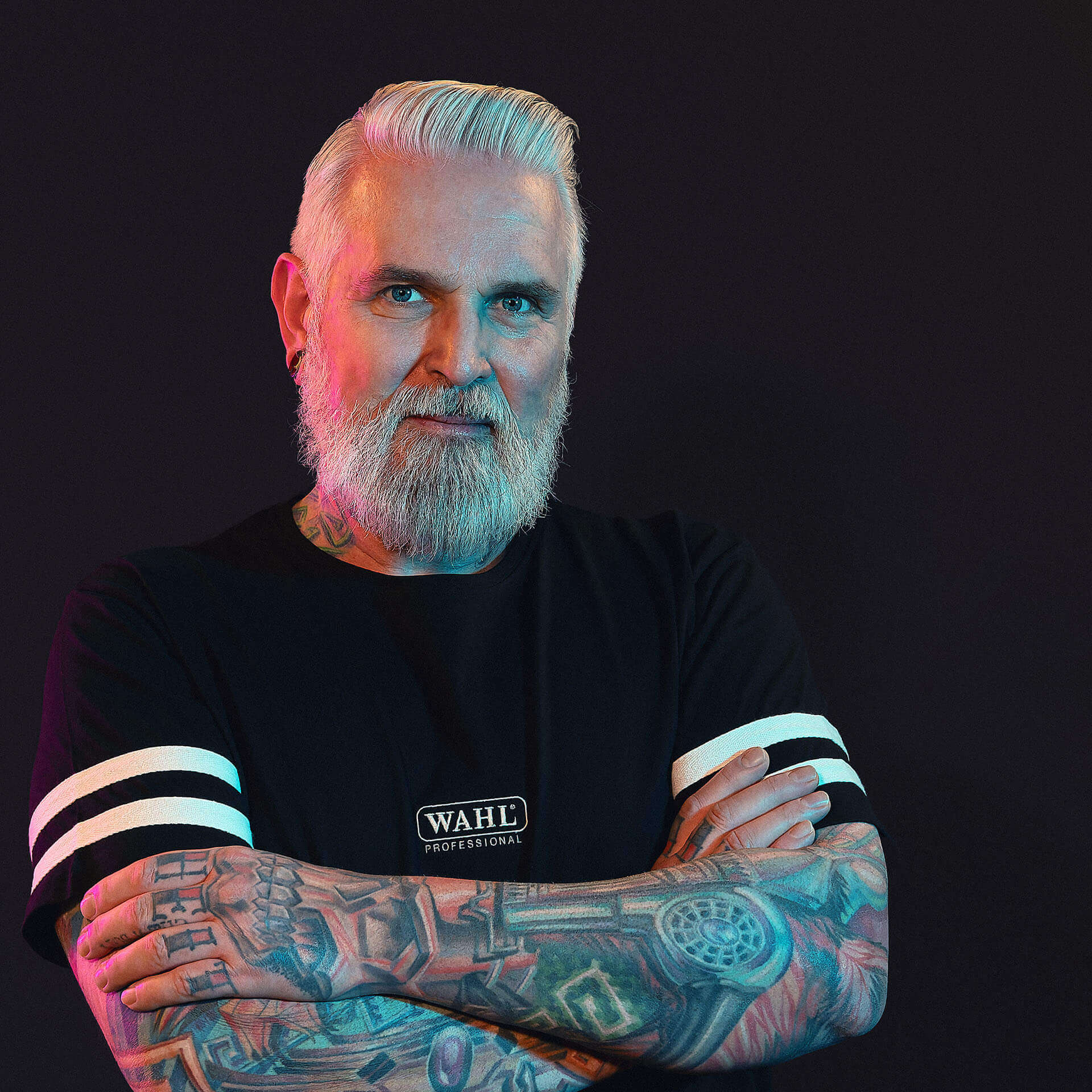 mann-mit-weissem-vollbart-und-tattoos-in-schwarzem-shirt-mit-verschraenkten-armen-vor-dunklem-hintergrund